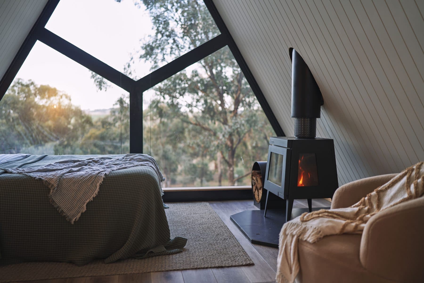 The wood heater going inside an A-Frame cabin, the bush through the gable window beyond