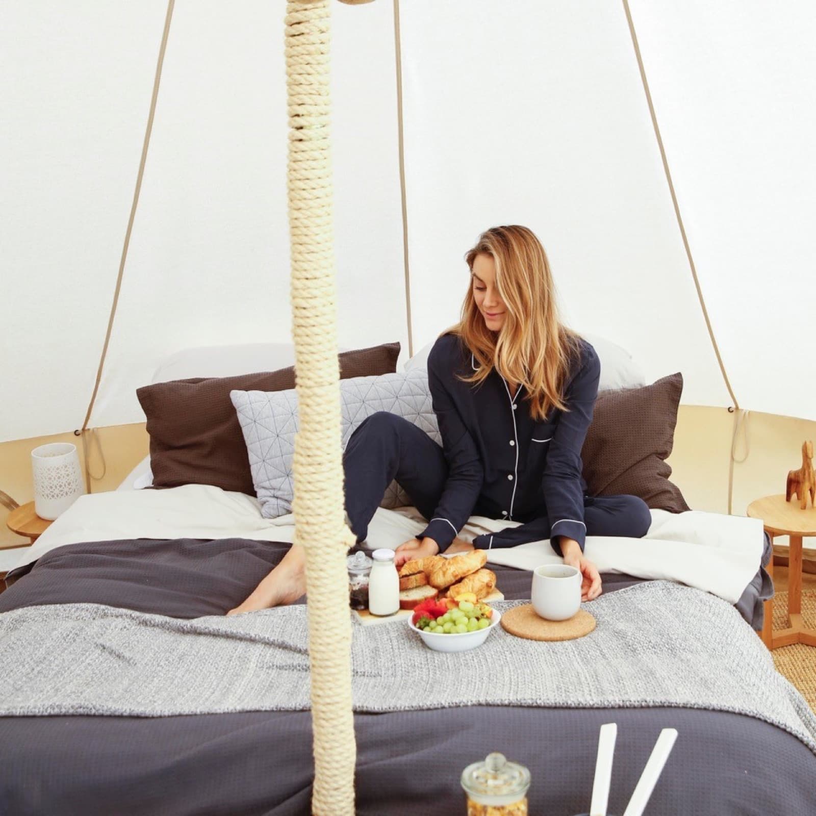 A guest sitting up in bed in a bell tent with the breakfast hamper unpacked around her