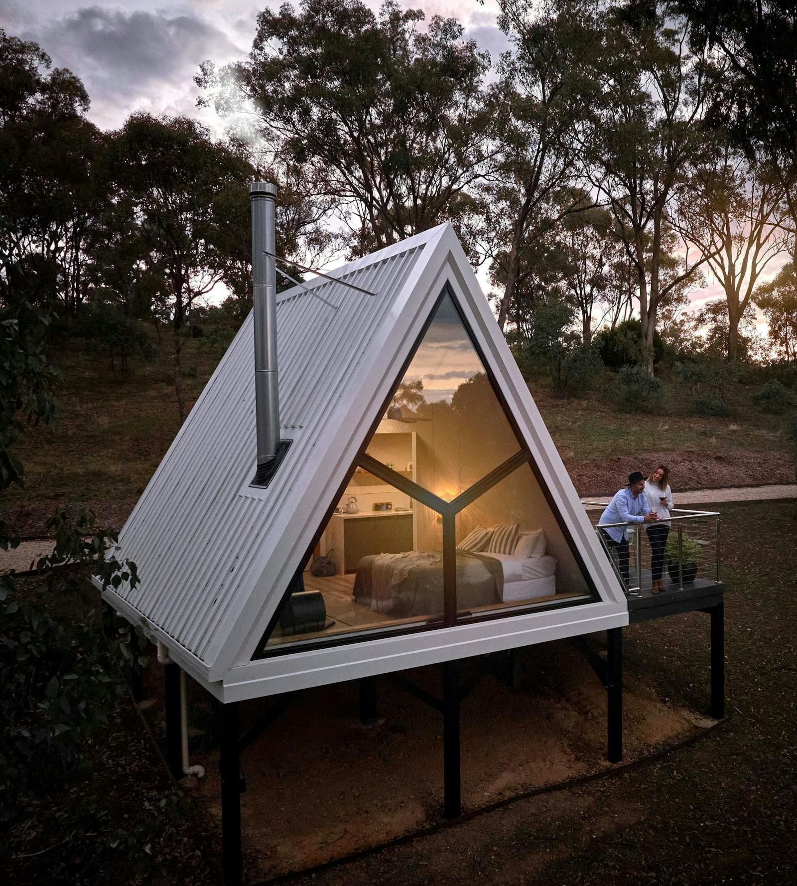 An A-Frame cabin at dusk, lit from inside, with guests sitting out on the deck