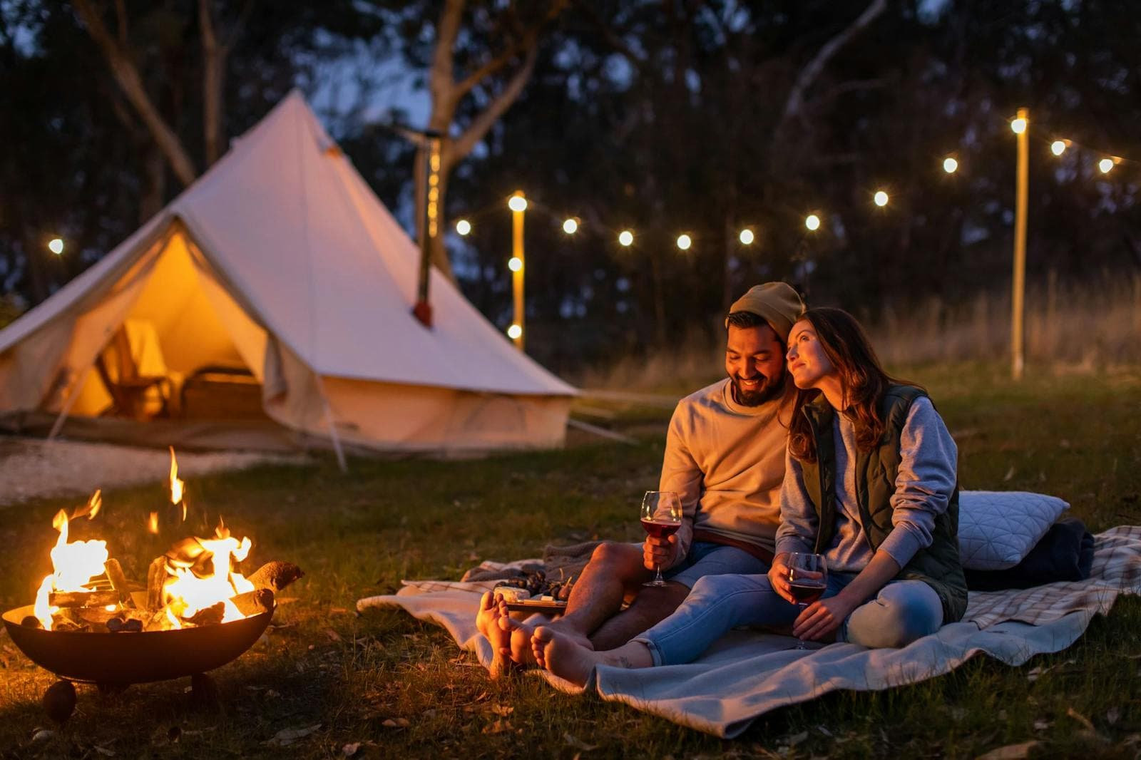 A couple by the fire pit at night, their bell tent glowing behind them under fairy lights