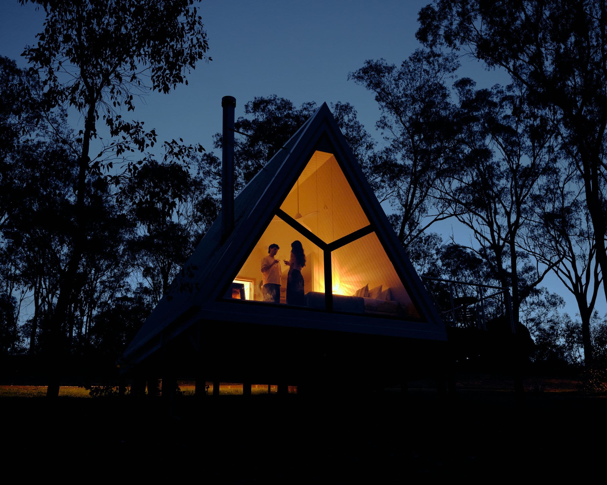 An A-Frame cabin at night, its glass gable glowing gold with two guests inside