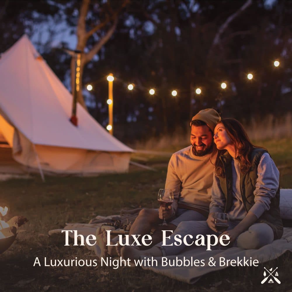 The Luxe Escape — a couple with wine by the fire under festoon lights beside a lit bell tent